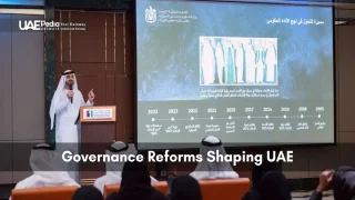 Man in kandura speaks at podium beside timeline slide while Emirati officials listen seated